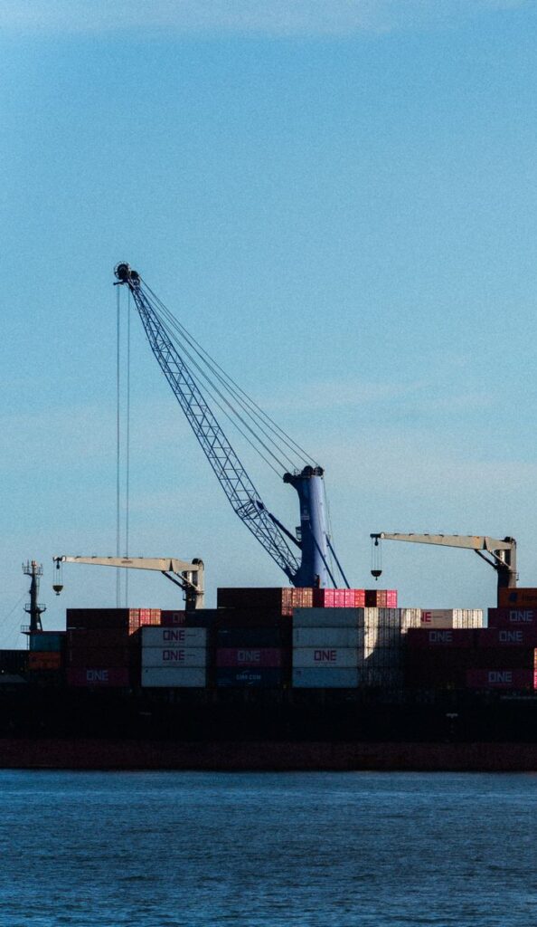 pexels-photo-38726115-38726115 View of a commercial shipping port with cranes and stacked cargo containers against a clear sky.